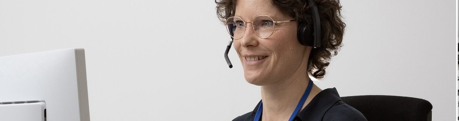 Woman with short curly hair wearing glasses and a headset, seated at a desk in front of a computer screen, smiling slightly.